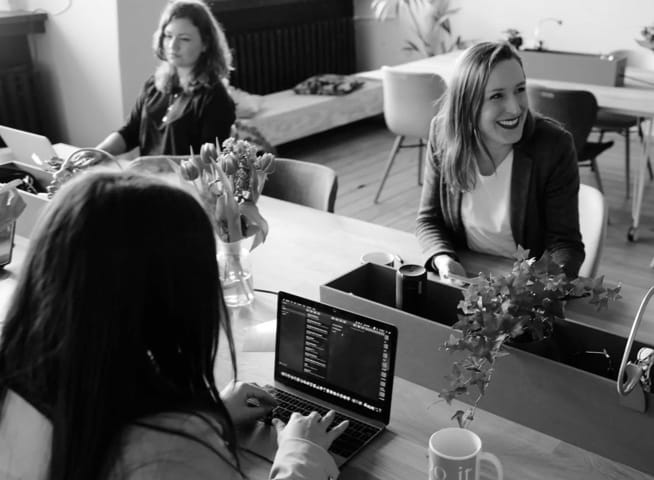 Three women seated at a conference table having a conversation while on their personal computers 